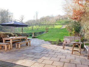 An outdoor patio with wooden benches and a table, surrounded by greenery and a stone pathway leading to a grassy area. - The Studio at Granny's Well (Mixtow, near Fowey)
