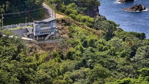 A coastal building with a winding road leading to it, surrounded by dense greenery and rocky outcrops.