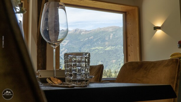 A dining table with a clear glass and a patterned glass mug, a wooden chair, and a view of mountains through a window. - Chalet ski hut holiday home (Flattach)