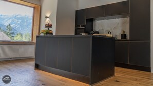 A modern kitchen with dark cabinetry, a central island, and a view of mountains through a large window. - Chalet ski hut holiday home (Flattach)