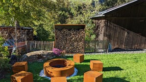 A garden with a circular fire pit, surrounded by six wooden stools, and a stack of firewood in the background. - Chalet ski hut holiday home (Flattach)