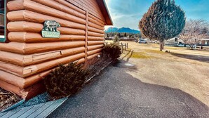 A log cabin with a 'Grizzly Lodge Welcome' sign, a paved driveway, and a view of a mountain range.