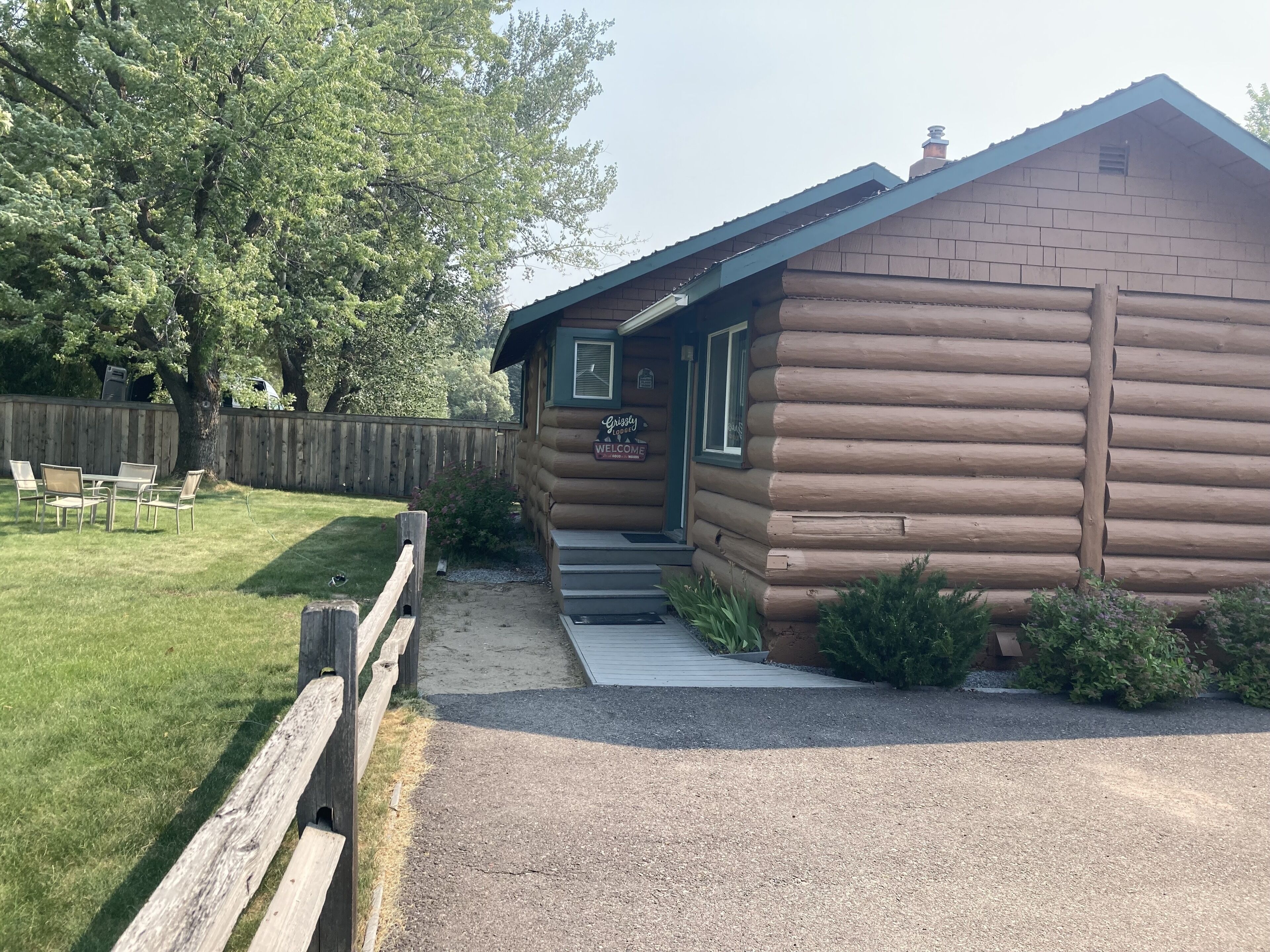 A log cabin with a 'Grizzly Lodge Welcome' sign, a gravel driveway, and a wooden fence.