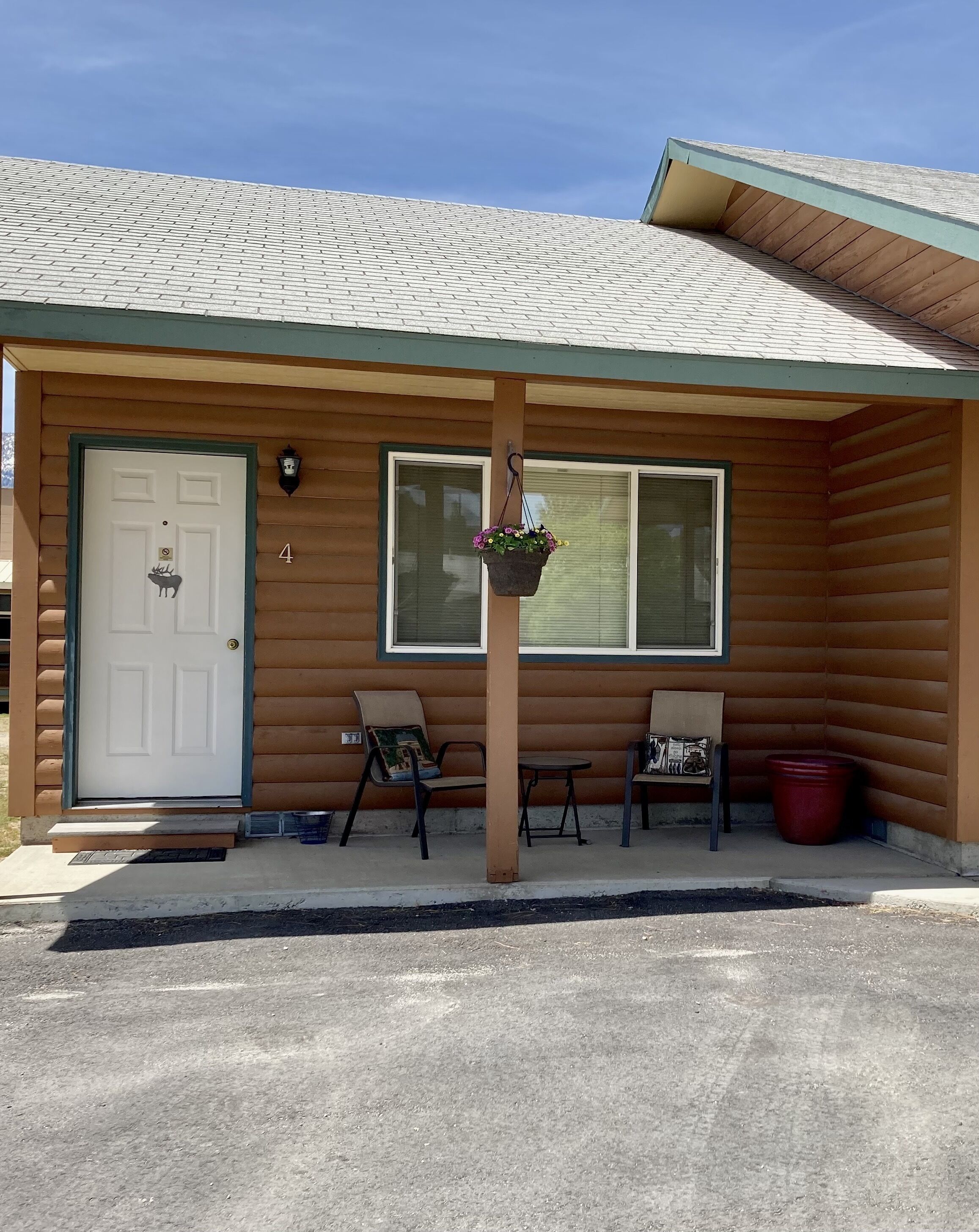 A log cabin with a white door, two windows, a porch with two chairs, and a hanging flower basket.