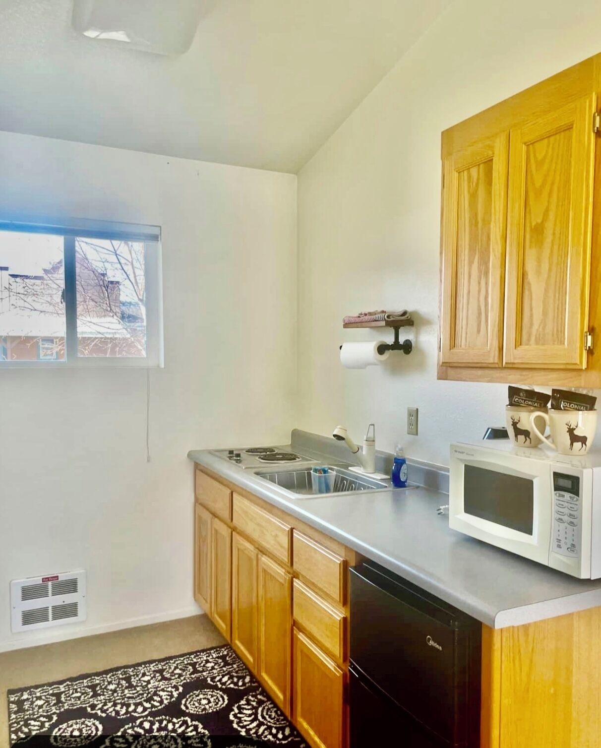A small kitchen with wooden cabinets, a stainless steel countertop, a microwave, a sink, and a window.