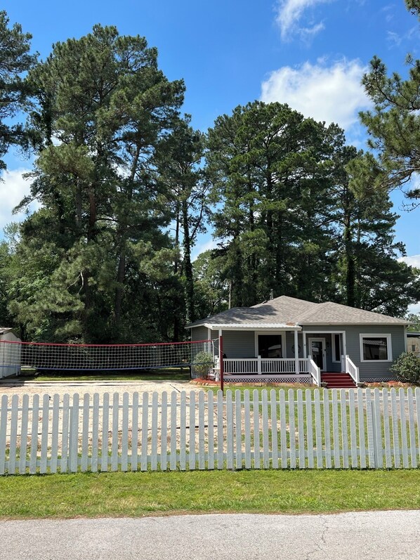 A single-story house with a porch, surrounded by a white picket fence and a red netted volleyball court.