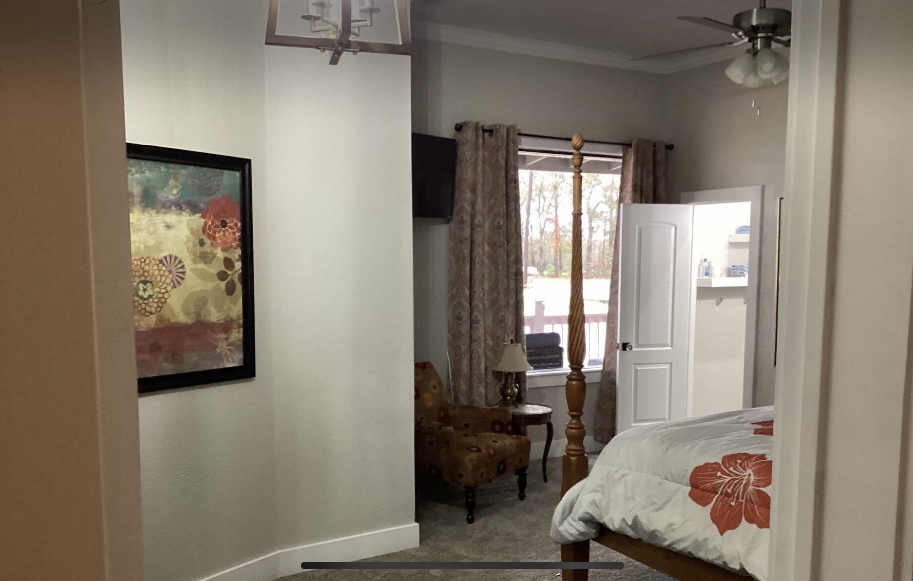 Un dormitorio con cama, una silla, un escritorio, un ventanal con cortinas y un cuadro en la pared.