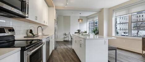 A modern kitchen with white cabinetry, a central island, and a view of the city through large windows.