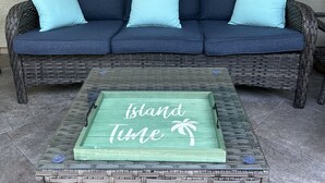 Een rieten bank met drie lichtblauwe kussens, een groen bijzettafeltje met de tekst 'Island Time' erop, en een gestructureerde muur op de achtergrond.