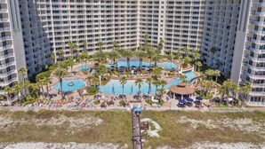 A high-rise hotel with multiple balconies, a central pool area surrounded by palm trees, and a beach access walkway.