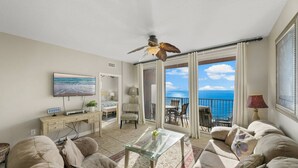 A spacious living room with a balcony overlooking the ocean, a ceiling fan, and a television on a desk.