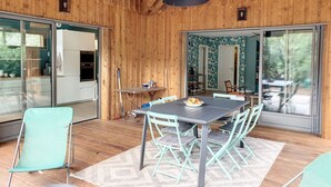 A wooden interior with a dining table and chairs, a kitchen area, and large sliding glass doors leading to an outdoor space. - 50 m from the lake, new wooden house - ideal family (Lacanau)