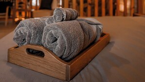 A wooden interior with a bed, a wooden tray with towels, and a stone wall in the background.