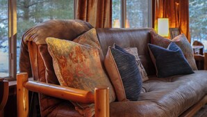 A log cabin interior with a leather sofa, wooden chair, and large windows with curtains.