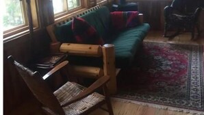 A cozy cabin interior with a wooden rocking chair, a green sofa with red cushions, and a large window providing natural light. - Cozy cottage trails, trout fishing & fireplace. (Marcell)