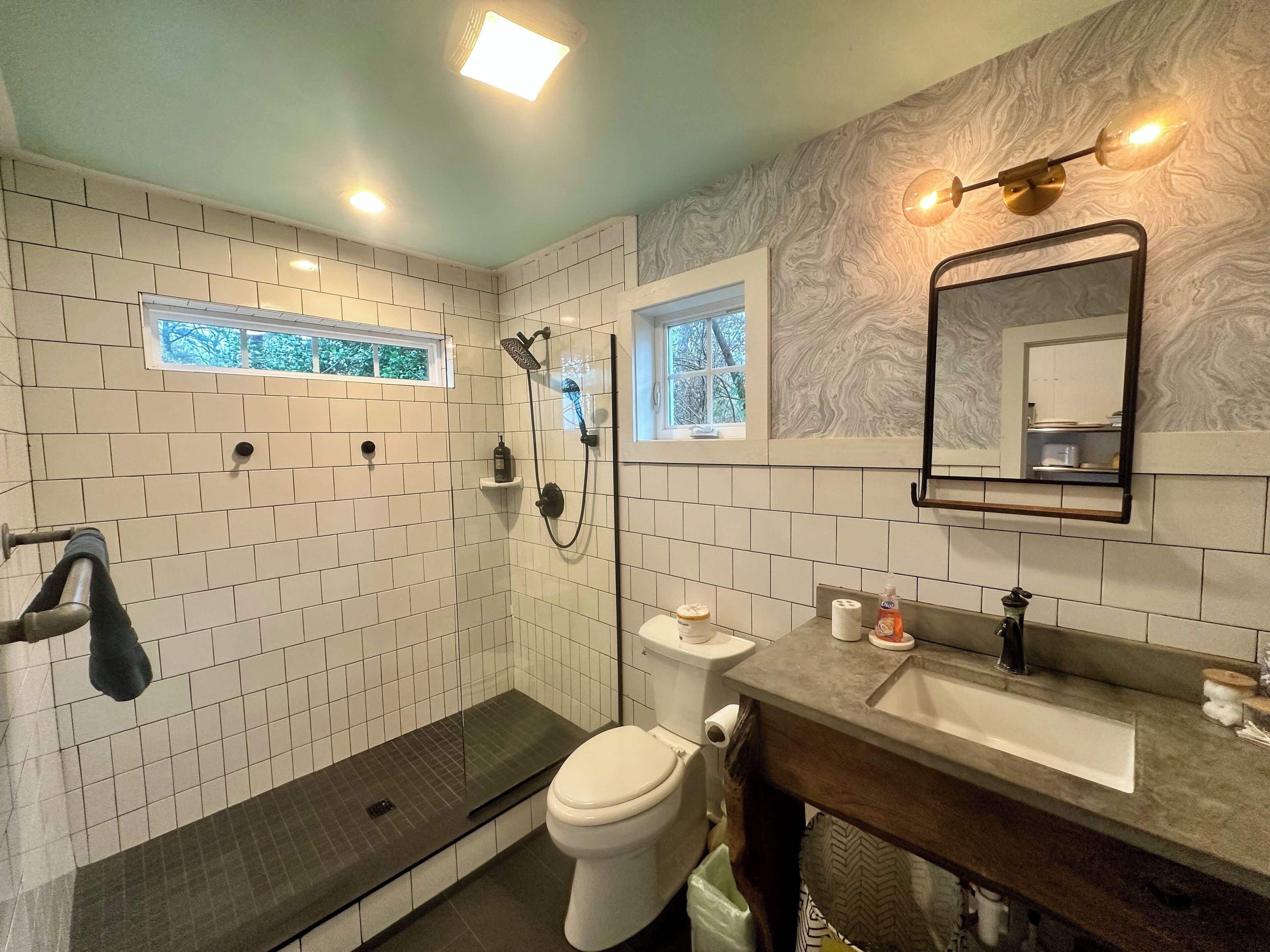 A bathroom with white subway tiles, a glass shower enclosure, a toilet, a sink with a mirror, and two windows.