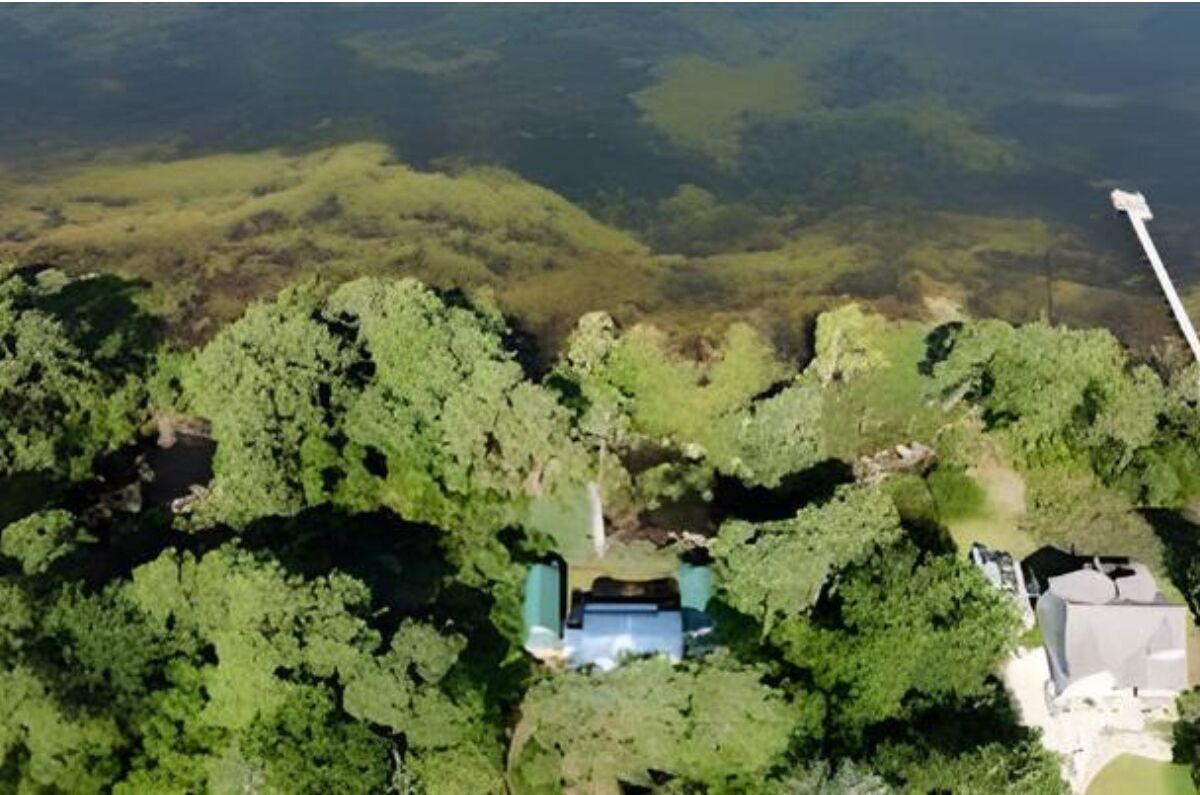 Luchtbeeld van een pand omringd door weelderige begroeiing, met een centraal gebouw met blauw dak, een wit gebouw aan de rechterkant en een steiger die in het water reikt.