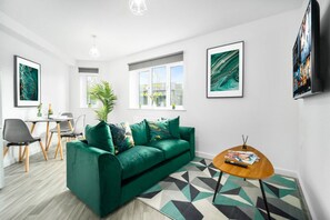 Ein modernes Wohnzimmer mit einem grünen Sofa, einem Couchtisch und einem Fernseher. An den Wänden hängen Bilderrahmen, und ein Fenster mit Jalousien ist zu sehen.