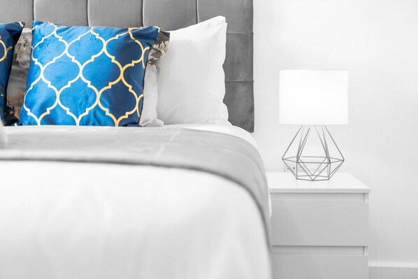 Una cama bien hecha con un almohadón blanco y azul estampado, un almohadón blanco liso y una lámpara geométrica sobre una mesita de noche blanca.