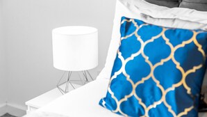 Ein ordentlich bezogenes Bett mit weißem Kopfteil, einem blau-goldenen Kissen und einer weißen Nachttischlampe auf einem weißen Nachttisch.