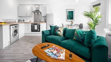 Ein modernes Wohnzimmer mit einem grünen Sofa, einem Couchtisch mit Zeitschriften, einer Waschmaschine und einer Küchenzeile mit weißen Schränken und Geräten.