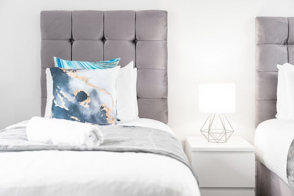 Una cama bien hecha con cabecera gris, ropa de cama blanca y un cojín decorativo. Hay una mesita de noche blanca con una lámpara geométrica al lado de la cama.