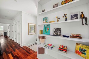 Een gang met witte muren en een houten vloer, die leidt naar een kamer met een witte boekenkast vol boeken en decoratieve objecten.