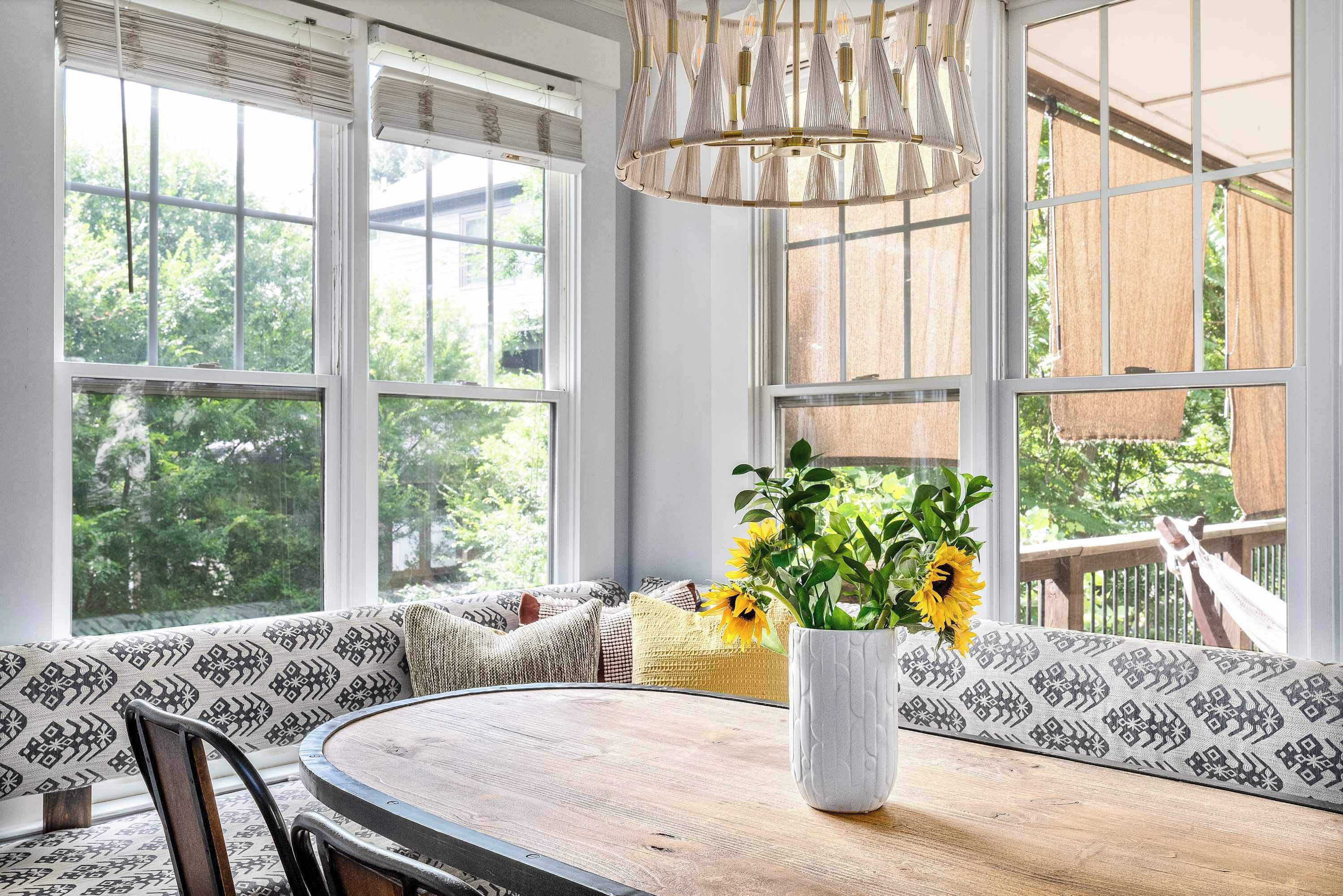 A dining area with a round table, a white vase with flowers, patterned cushions, and large windows with a view of greenery.