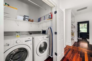 Un cuarto de lavandería con dos lavadoras de carga frontal, repisas con contenedores de almacenamiento y una puerta que da a otra habitación.
