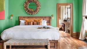 Un dormitorio con cabecera de madera, una cama grande con ropa de cama blanca, un banco tejido y una pared verde con un espejo circular de madera.