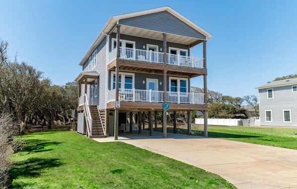 A two-story house with a balcony on the upper floor, a covered patio area, and a front yard. - DownWinder- Spacious 3 BR House - Walk to Beach (Kitty Hawk)