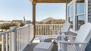 A wooden deck with white railings and Adirondack chairs, overlooking a residential area and a hill. - DownWinder- Spacious 3 BR House - Walk to Beach (Kitty Hawk)