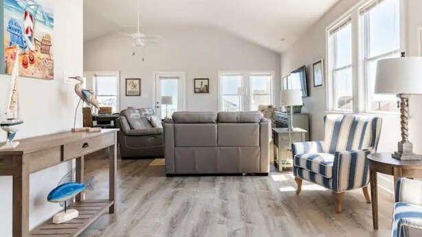 A spacious living room with a gray sofa, striped armchair, and wooden coffee table. There are large windows providing natural light and a ceiling fan. - DownWinder- Spacious 3 BR House - Walk to Beach (Kitty Hawk)