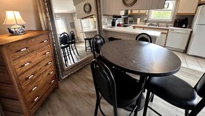 A kitchen with a round table and chairs, a large mirror, wooden cabinets, and a hanging light fixture. - KH Escape! Pool+Tennis+Basketball+0.8 mi to Beach! (Kitty Hawk)
