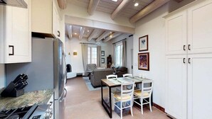 Una cocina con refrigerador, estufa y mesa de comedor con sillas. Se observa un sofá y una ventana al fondo.