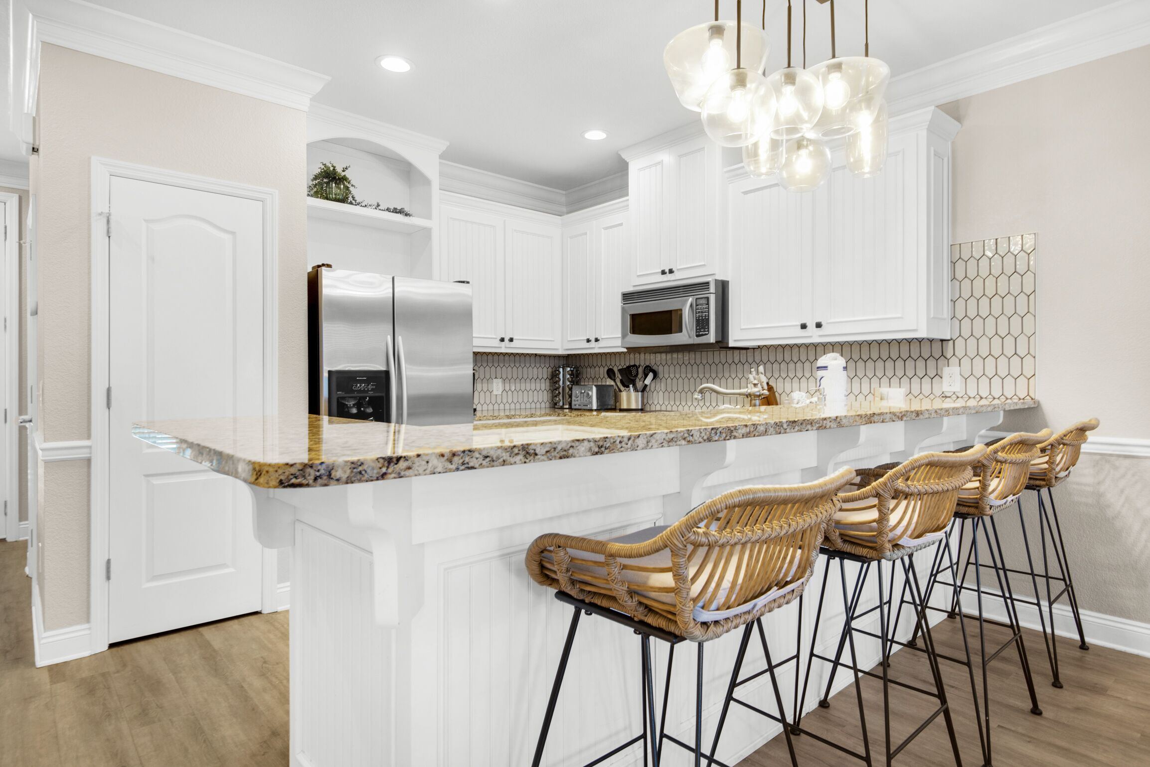 A modern kitchen with white cabinetry, a granite countertop, stainless steel appliances, and a bar area with wicker stools.