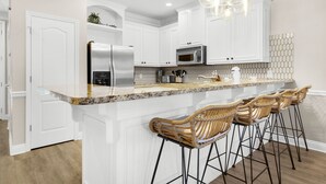 A modern kitchen with white cabinetry, a granite countertop, stainless steel appliances, and a bar area with wicker stools.