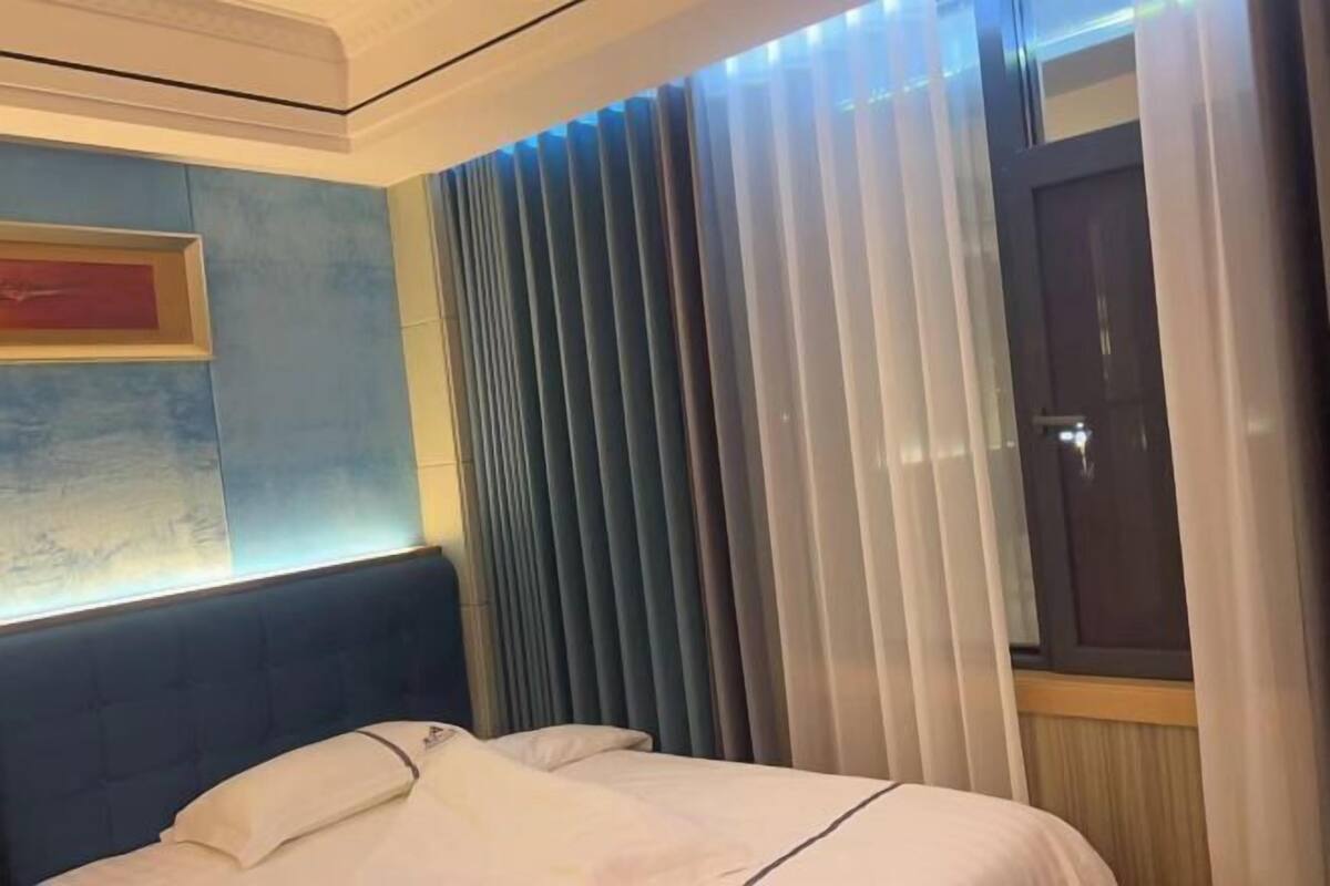 A hotel room with a bed, a headboard, a window with blinds, and a door.