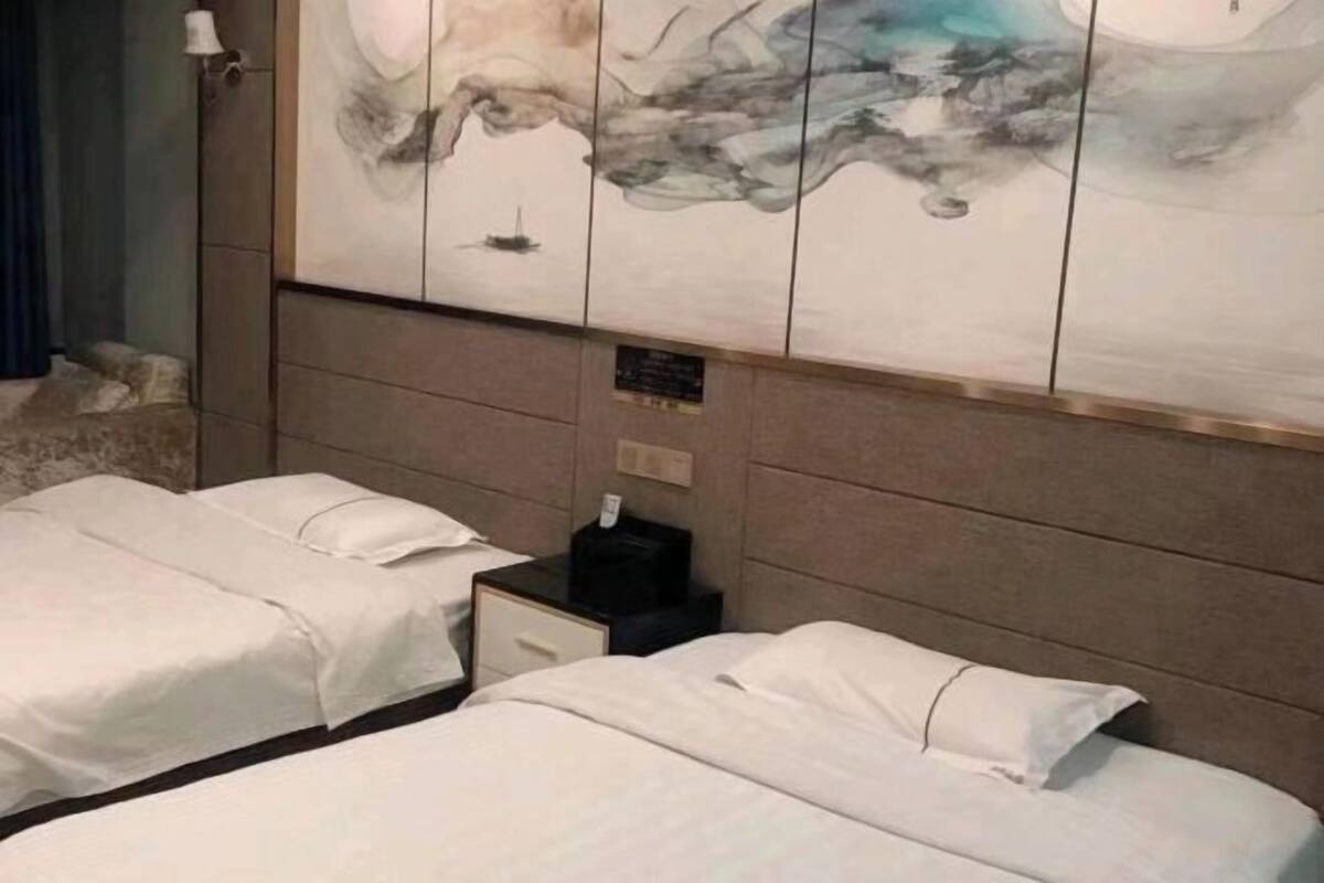 A hotel room with two beds, a nightstand, a wall-mounted artwork, and a small table with a lamp.