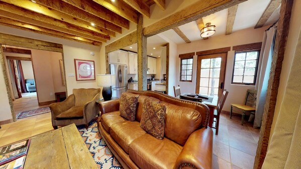 A spacious living room with a leather sofa, wooden coffee table, and a patterned rug. A kitchen is visible in the background with wooden cabinets and a dining area.