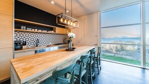 Cozinha moderna com bancada de madeira, banquetas e vista para montanhas através de amplas janelas.