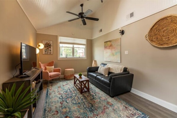 A living room with a black leather sofa, a pink armchair, a wooden coffee table, a television, and a ceiling fan.