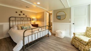 Un dormitorio con estructura de cama metálica, lámpara de noche, espejo redondo en la pared y sillón estampado.