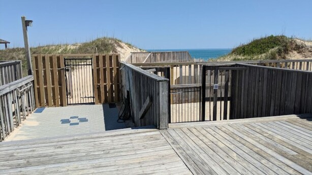 Un paseo marítimo de madera que conduce a una zona de playa con un sendero arenoso y una valla de madera.