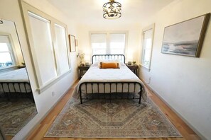 A bedroom with a bed, a rug, a chandelier, and a painting on the wall.