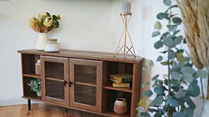 A wooden cabinet with glass doors, a vase with dried flowers, and a tall candle holder on top.