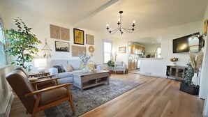 A spacious living room with a wooden floor, a sofa, a coffee table, and a chair. There is a large plant and artwork on the walls.