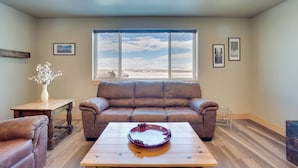 A spacious living room with a brown leather couch, a wooden coffee table, and a large window offering a view of the sky and landscape. - Saratoga Retreat ~ 3 Mi to Saratoga Lake! (Saratoga)