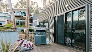 Een modern terras buiten, met een barbecuegrill en gasfles, omgeven door een houten terras en een glazen schuifdeur die leidt naar een overdekt gedeelte.