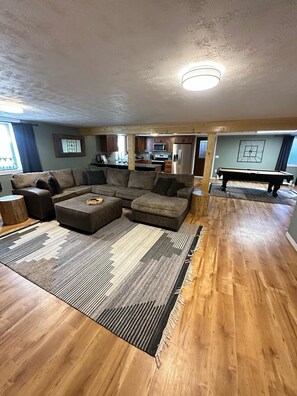 Ein geräumiges Wohnzimmer mit einer großen Eckcouch, einem Couchtisch und einem Billardtisch. Der Raum hat Holzböden und im Hintergrund ist eine Küchenzeile zu sehen.